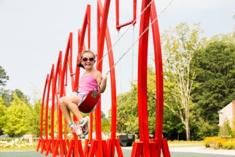 The red artistic swing set and lush trees at Abernathy Greenway Playable Art Park